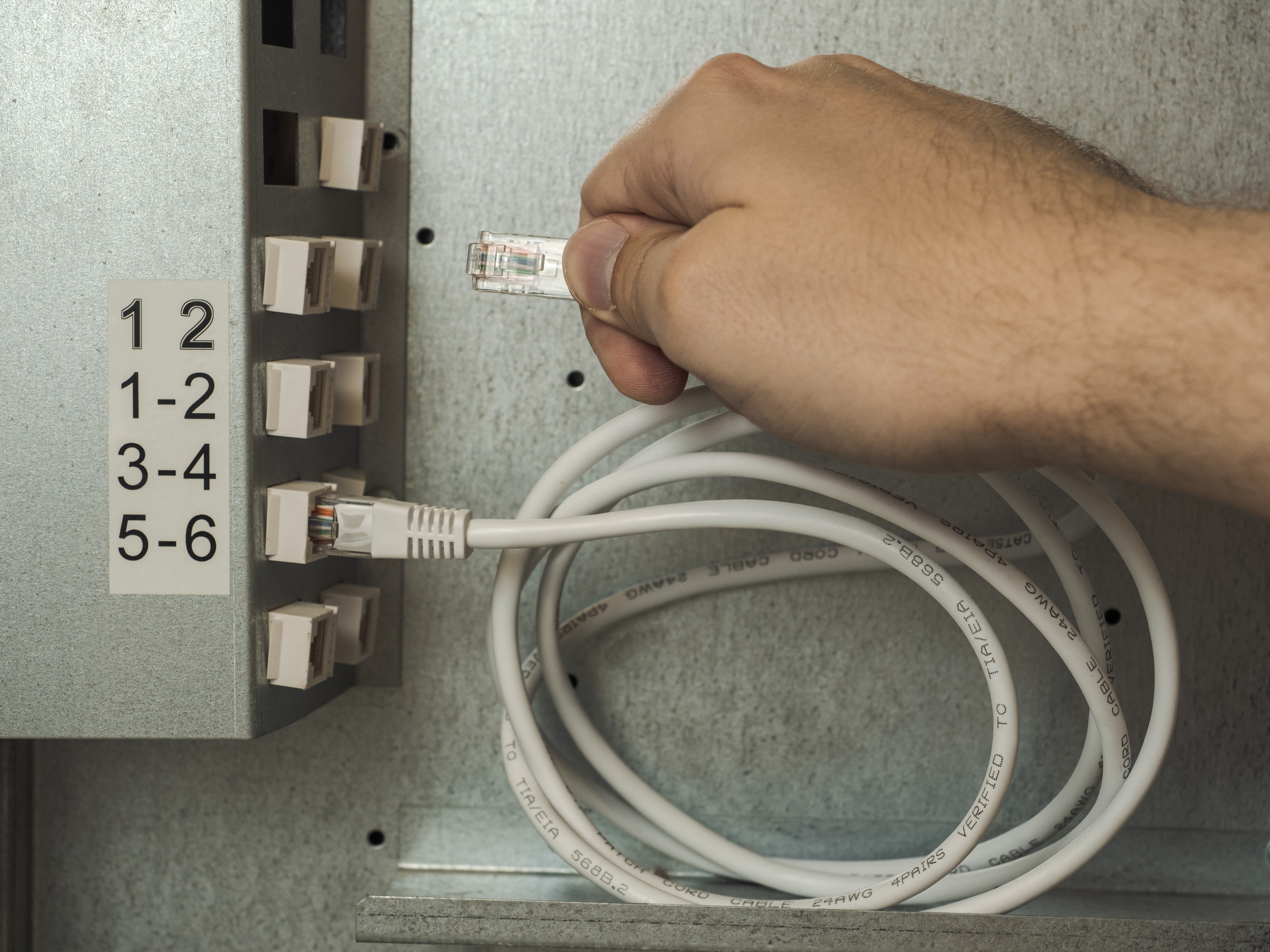 Close-up of a technician's hand plugging a white RJ45 ethernet patch cord cable into a network patch panel socket inside a server cabinet. High-speed internet connection, local area network (LAN), and IT infrastructure concept.
